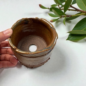 May include: Handmade ceramic planter with a brown and tan glaze. The small, round pot has a drainage hole in the bottom and a textured, uneven rim. The planter is suitable for small plants and succulents.