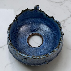 May include: A handmade, blue ceramic bowl with a unique, irregular rim and a central hole. The bowl has a textured surface and a dark brown rim, creating a rustic aesthetic. This decorative piece is ideal for home decor.