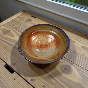 May include: A handmade ceramic bowl with a spiral design in shades of orange, tan, and brown. The rim is a dark blue-purple colour. The bowl is on a wooden surface.