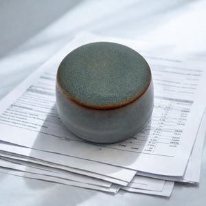 May include: A small, rounded ceramic cup with a matte, teal-green exterior and a warm brown rim. The cup sits atop a stack of white papers, suggesting a workspace or office setting. The cup's design is simple and modern.