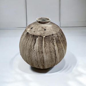 May include: A handcrafted, textured ceramic vase with a bulbous body and a narrow neck. The vase features a mottled, earthy color palette of gray and brown, with subtle vertical ridges and dark spots. The opening is a light beige color.