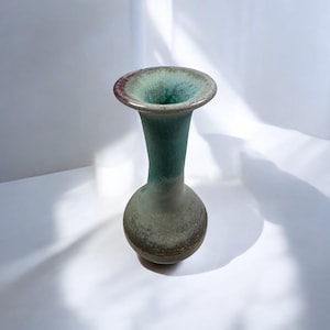 May include: A ceramic vase with a wide, flared rim and a rounded base. The vase has a gradient of colors, transitioning from a dark green at the top to a lighter green and gray at the base. The rim has a reddish-brown accent.