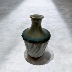 May include: A ceramic vase with a distinctive design. The vase features a narrow neck and a wider body with a textured, swirling pattern. The colour is a blend of grey, green, and teal, with a dark teal rim. The vase casts a shadow.