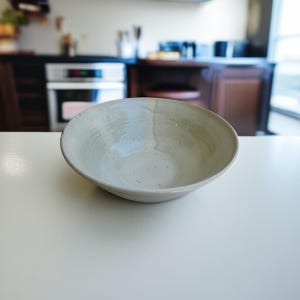 Może przedstawiać: Ręcznie robiona, kremowa ceramiczna miska o szerokim, płytkim kształcie. Miska ma lekko nieregularny kształt i teksturowaną powierzchnię, co sugeruje rustykalną estetykę. Miska stoi na białej powierzchni.