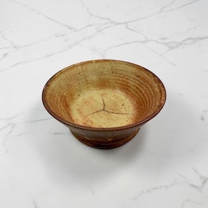 May include: A handmade ceramic bowl with a pedestal base. The bowl has a light brown interior with a cracked pattern and a darker brown exterior. The bowl's rim and base are a darker shade of brown.
