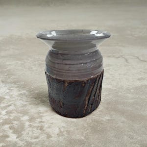 Puede incluir: Un jarrón de cerámica hecho a mano con una base texturizada de color marrón oscuro y una sección superior lisa de color gris. El jarrón tiene un borde acampanado y una forma cilíndrica, mostrando una estética rústica. El jarrón es adecuado para exhibir flores o como objeto decorativo.
