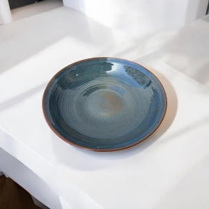 Puede incluir: Un plato redondo de cerámica con un esmalte azul y marrón. El plato tiene un borde ancho y una superficie ligeramente texturizada. Los colores se mezclan, creando un aspecto único y artístico. El plato es adecuado para servir alimentos.