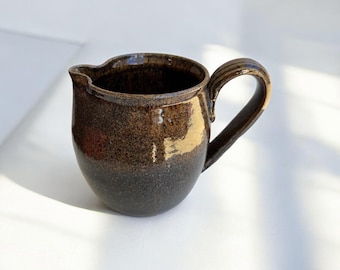 May include: A handmade ceramic creamer with a dark brown glaze. The creamer has a rounded body, a spout for pouring, and a curved handle. The glaze transitions from a lighter brown at the top to a darker brown at the base. The creamer is designed for serving liquids.