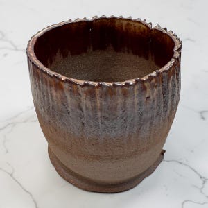 May include: A handmade ceramic vessel with a textured, ridged exterior and a dark brown, glossy interior. The rim has a jagged, uneven edge. The color gradient transitions from a light brown at the base to a darker, almost black, hue at the top.