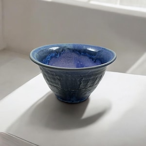 May include: A handcrafted ceramic bowl with a textured exterior and a glossy, deep blue interior. The bowl has a wide rim and a rounded shape, suitable for serving or display. The bowl is approximately 15 cm in diameter and 10 cm tall.