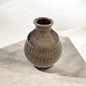 May include: A small, handcrafted ceramic vase with a textured, ribbed design around the middle. The vase has a rounded body and a narrow neck with a dark burgundy interior. The exterior is a muted olive green with subtle color variations.