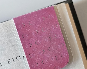 May include: A pink bookmark with a silver geometric pattern. The bookmark is made of a textured material and has a clear plastic cover.