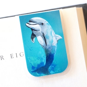 May include: A blue and white dolphin bookmark with a watercolor effect. The dolphin is jumping out of the water with its tail in the air. The background is a light blue with white and green watercolor splashes.