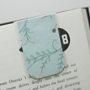 Könnte beinhalten: Ein hellblaues Lesezeichen mit einem weißen und hellblauen Blumenmuster. Das Lesezeichen ist an einem Buch mit einem schwarzen Seitenmarker befestigt, auf dem "8" steht.