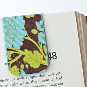 Könnte beinhalten: Ein dekorativer Lesezeichen mit blauem Rand, verziert mit einem Blumenmuster in Blau- und Grüntönen auf dunklem braunem Hintergrund. Das Lesezeichen befindet sich in einem Buch, wobei die Seitenzahl 48 sichtbar ist.