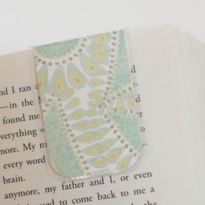 Könnte beinhalten: Nahaufnahme eines teal- und weiß gemusterten Lesezeichens mit einer transparenten Plastikhülle. Das Lesezeichen liegt auf einem Buch mit Text.