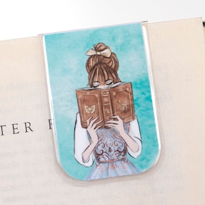 May include: A clear, U-shaped bookmark featuring an illustration of a woman reading a book. The woman has brown hair and is wearing a blue dress. The book is brown with a butterfly and insect design. The background is a teal color.