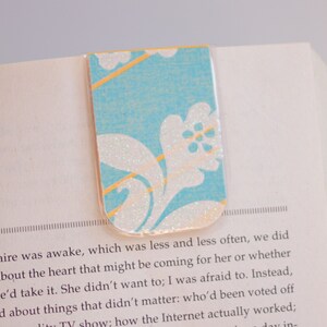 Könnte beinhalten: Ein blaues und weißes Blumen-Lesezeichen mit goldenen Akzenten. Das Lesezeichen ist aus Papier und hat eine glitzernde Oberfläche.