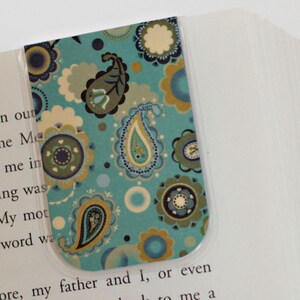 Könnte beinhalten: Ein dekorativer Lesezeichen mit einem türkisfarbenen Paisley-Muster und floralen Akzenten in Blau, Grün und Gold. Das Lesezeichen befindet sich in einem Buch mit weißen Seiten und einem blauen Buchrücken.