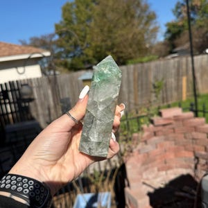 May include: A clear, rectangular crystal with green and grey hues. The crystal is held up against a bright blue sky. The crystal is a natural, polished stone.