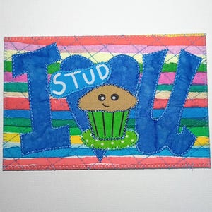 May include: A colorful, handmade fabric art piece with the word "I LOVE STUD" in blue letters. A cupcake with a green liner and a smiling face is in the center. The background is striped with various colors.