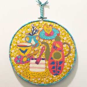 May include: A round, yellow fabric wall hanging with a blue border. The fabric features a colorful embroidered design of a teapot, teacups, a cake, and a bird. The design is surrounded by a yellow floral pattern.