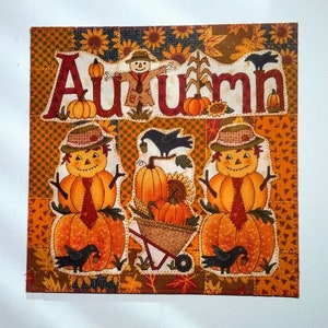 May include: A square patchwork design with the word "Autumn" in large letters, featuring pumpkins, scarecrows, and crows. The background is a mix of orange, brown, and yellow fabrics with a stitched border. Sunflowers and corn stalks are also present.