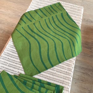 May include: Green cloth napkins with a wavy dark green stripe design. The napkins are folded and arranged on a white wicker surface. The design is repeated on the other napkins.