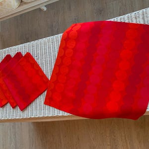 May include: A set of red and orange fabric napkins and a matching tablecloth. The fabric features a repeating pattern of large circles in varying shades of red. The items are displayed on a white wicker surface.
