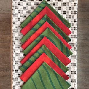 May include: A collection of folded cloth napkins in alternating red and green. The red napkins feature a circular pattern, while the green napkins have a striped design. The napkins are arranged on a white wicker surface.