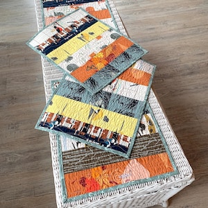 May include: A quilted table runner and two matching placemats. The runner and placemats feature a patchwork design with various fabrics in shades of orange, yellow, blue, and brown. The placemats are square and the runner is rectangular.