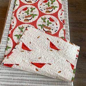 May include: A quilted table runner and placemat set featuring a cardinal bird design. The runner has a red and white pattern with circular designs. The placemat has a white background with red cardinal birds and leaf accents. The set is placed on a white wicker table.