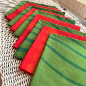 May include: A collection of folded cloth napkins in a fan arrangement. The napkins feature various patterns, including stripes and floral designs, in shades of red, orange, and green. The napkins are arranged on a white wicker surface.