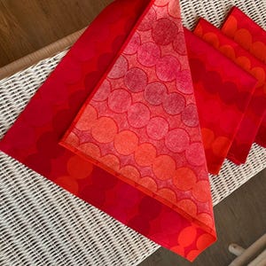 May include: A set of red and orange patterned table linens. The design features overlapping circles in varying shades of red and orange. The linens are arranged on a white wicker surface.