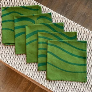 May include: A set of six green cloth napkins with a wavy, dark green pattern. The napkins are neatly folded and arranged on a white wicker surface, creating a visually appealing display. The color palette is dominated by shades of green.