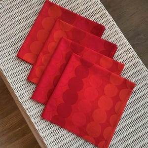 May include: Four red cloth napkins with a repeating pattern of large circles in varying shades of red and orange. The napkins are neatly arranged on a white wicker surface, creating a vibrant and eye-catching display.
