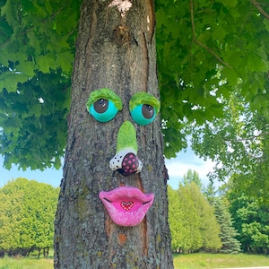 May include: A whimsical tree face sculpture with turquoise eyes, a green nose with polka dots, and pink lips with a heart. The sculpture is attached to a tree trunk, creating a playful outdoor art piece.