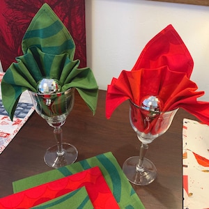 May include: Two wine glasses with folded green and red napkins, each holding a small disco ball. Additional green and red napkins are visible on the table. The napkins are folded in a decorative style, suggesting a festive or holiday theme.