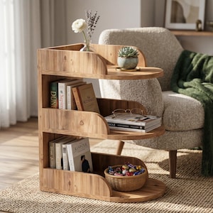 Puede incluir: Una mesa auxiliar de madera de tres niveles con un marco curvo en forma de S. Los estantes contienen varios libros, incluidos Sapiens y The Alchemist, una pequeña suculenta, un jarrón de vidrio con flores, gafas de lectura y una cesta tejida.