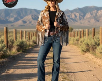 May include: A woman wearing a tan cowboy hat, a brown and white cowhide jacket with fringe, a black top, blue jeans, and brown boots. The background features a dirt path, wooden fence posts, and mountains.