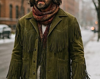 May include: A man wearing a brown fedora hat, a fringed olive green suede jacket, and a patterned scarf. He is also wearing blue jeans. The background shows a snowy street and buildings.