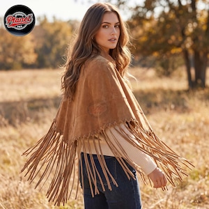 Women's genuine suede leather fringe western boho cape poncho — A tan suede poncho featuring a whip-stitched hemline with long, dangling fringe trim. The garment is worn over a cream-colored ribbed knit sweater and dark blue denim jeans, showcasing a