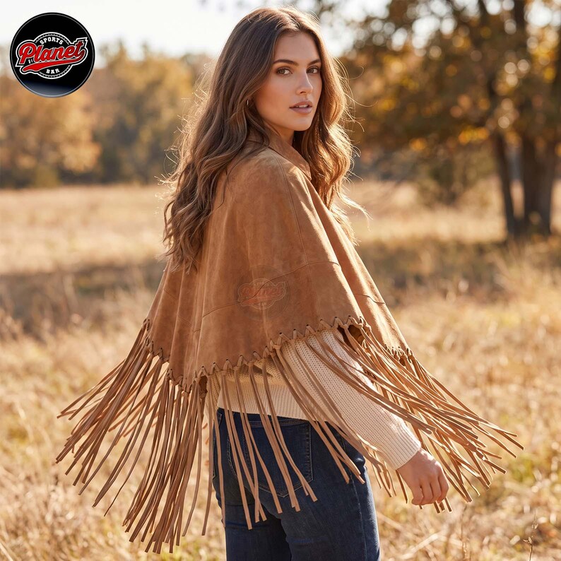 Women's genuine suede leather fringe western boho cape poncho — A tan suede poncho featuring a whip-stitched hemline with long, dangling fringe trim. The garment is worn over a cream-colored ribbed knit sweater and dark blue denim jeans, showcasing a