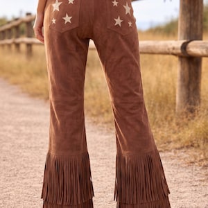 Puede incluir: Pantalones de ante marrón con detalles de estrellas blancas en los bolsillos traseros y flecos en el dobladillo. Los pantalones tienen un diseño de pierna acampanada, creando un look de inspiración occidental. Los pantalones son de color marrón medio.