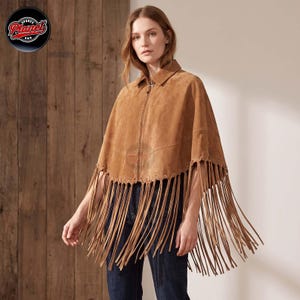 Women's genuine suede leather fringe western boho cape poncho — A tan suede poncho featuring a full-length front zipper, a classic collar, and long fringe detailing along the hemline. The garment is worn over dark blue denim jeans, showcasing a textu
