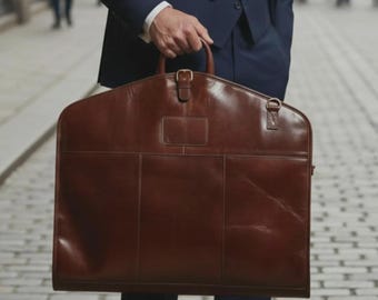 Könnte beinhalten: Eine braune Leder-Kleidersack mit Griff und Schnallenverschluss. Die Tasche hat eine rechteckige Form mit gewölbtem Oberteil. Die Tasche wird von einer Person in einem marineblauen Anzug gehalten. Klassisches, professionelles Design.
