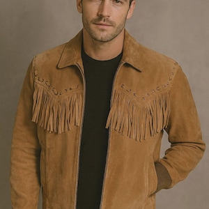May include: A tan suede jacket with fringe detailing across the chest and sleeves. The jacket has a collared neckline, a zip closure, and ribbed cuffs. The model is wearing a black shirt underneath.