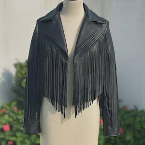 May include: Black leather jacket with fringe detailing. The jacket has a classic collar and long sleeves. The fringe is along the front and sleeves. The jacket is displayed on a mannequin.