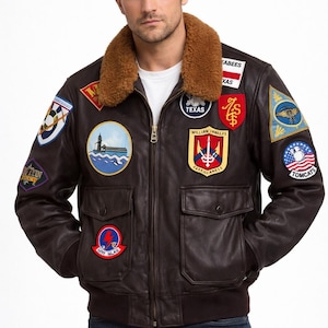 Peut inclure: Veste de bombardier en cuir marron avec plusieurs écussons, un col en fourrure marron et deux poches avant. La veste a une fermeture éclair et un ourlet côtelé. Les écussons incluent les mots "Texas" et "Tomcats".