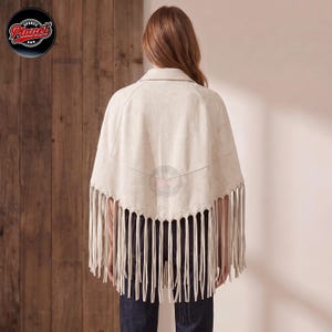 Women's genuine suede leather fringe western boho cape poncho — A beige suede-like poncho featuring a pointed hemline adorned with long, thin fringe detailing. The garment has a structured collar and a V-shaped seam across the back. The fabric has a